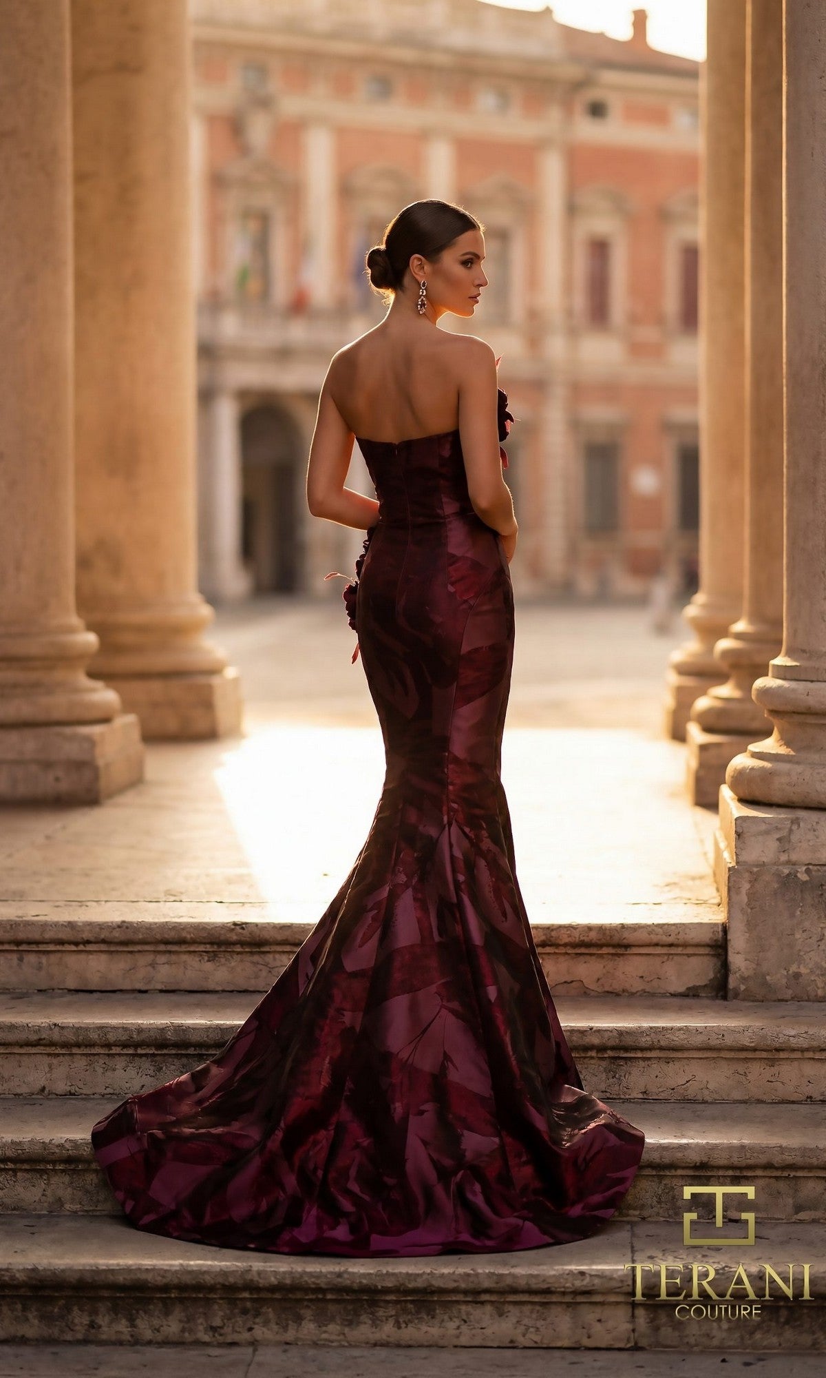 A woman in the Terani 261E6259 long formal dress with a strapless mermaid silhouette stands on stone steps framed by columns and a grand building. The ?TERANI COUTURE? logo is displayed at the bottom right.